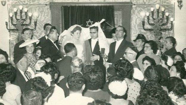 والتقط هذه الصورة أحد ضيوف حفل زفاف هذا العريس، غابي تمام، على عروسه لينا إيليني في المعبد عام 1958، ويظهر فيها الحاخام الباز وهو يتلو مباركته ممسكا بكأس من النبيذ.